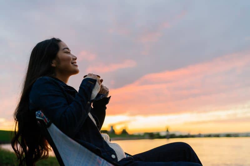 A young woman is enjoying sitting near a lake and drinking a hot drink in nature during sunset. A young woman is enjoying sitting near a lake and drinking a hot drink in nature during sunset.