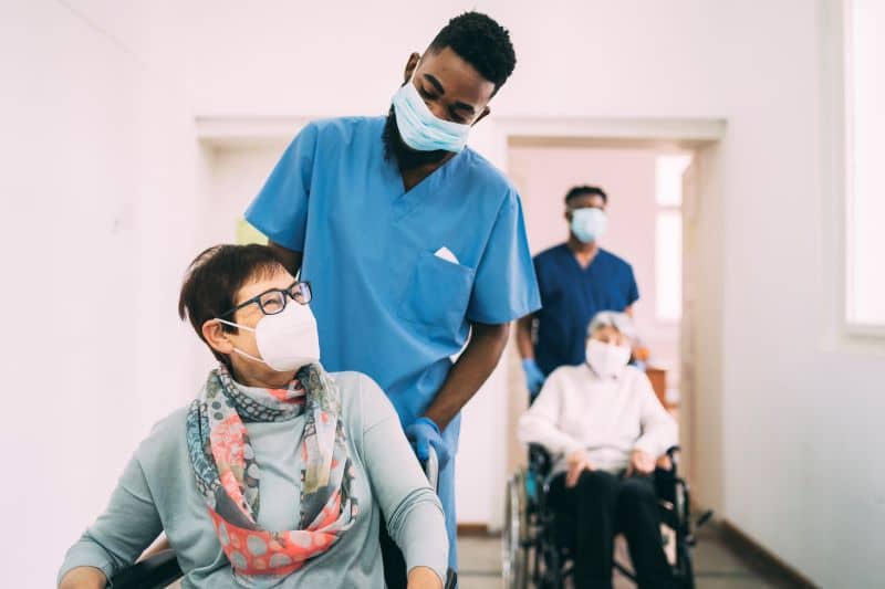 Two senior female patients in wheelchairs, being pushed by male healthcare workers along a corridor, all are wearing protective face masks Two senior female patients in wheelchairs, being pushed by male healthcare workers along a corridor, all are wearing protective face masks